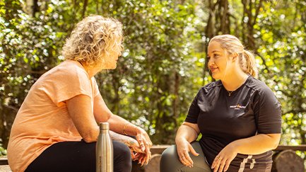 Mates4Mates staff member kneeling down talking with a female client who is sitting on a log outdoors with trees in the background. 