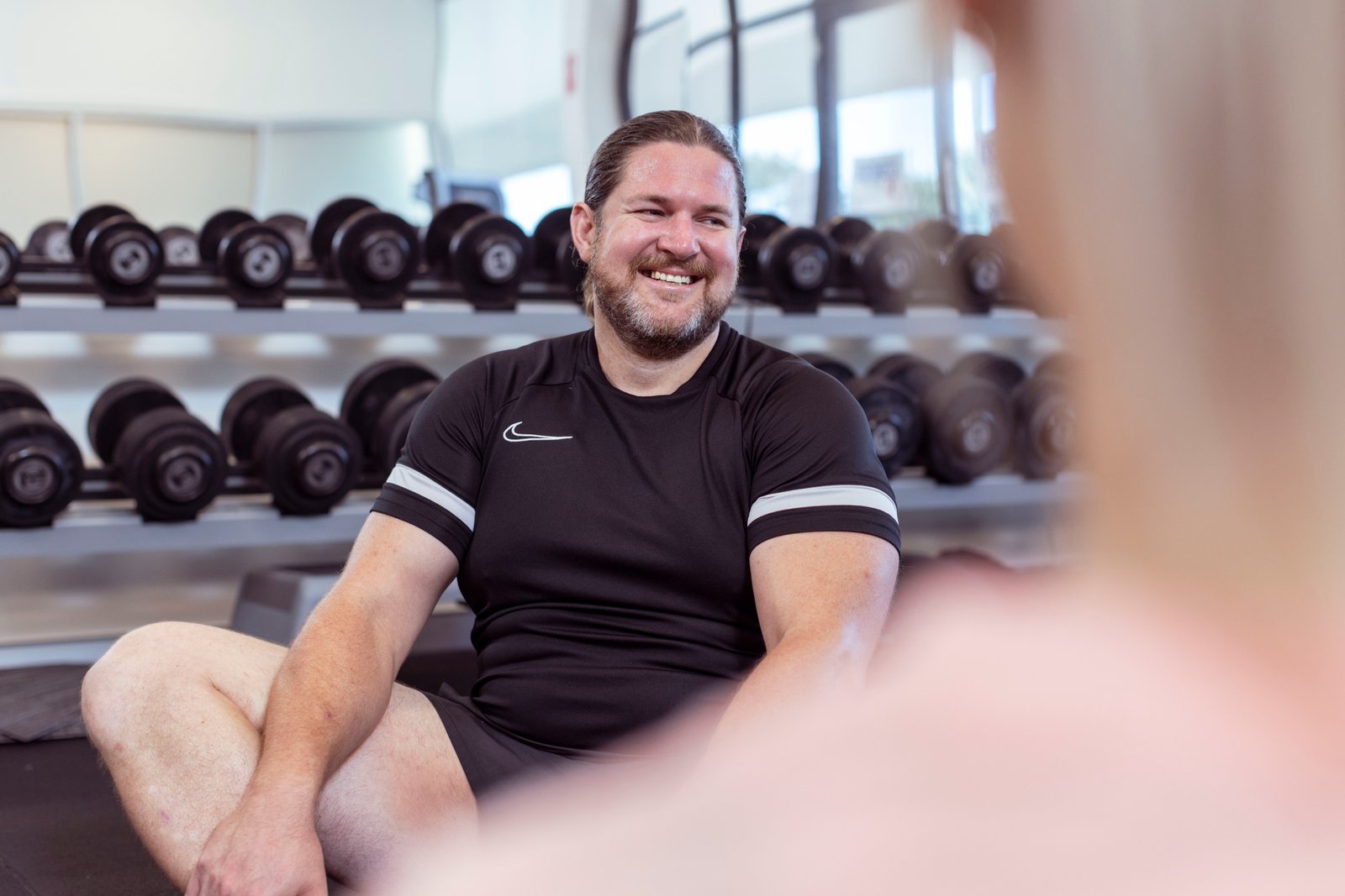 Man sitting on the floor of a gym in front of a rack of dumbbells, smiling at others sitting with him in the gym. 
