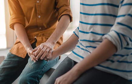 Two people sitting beside each other, one consoling the other with hands placed on the others. 